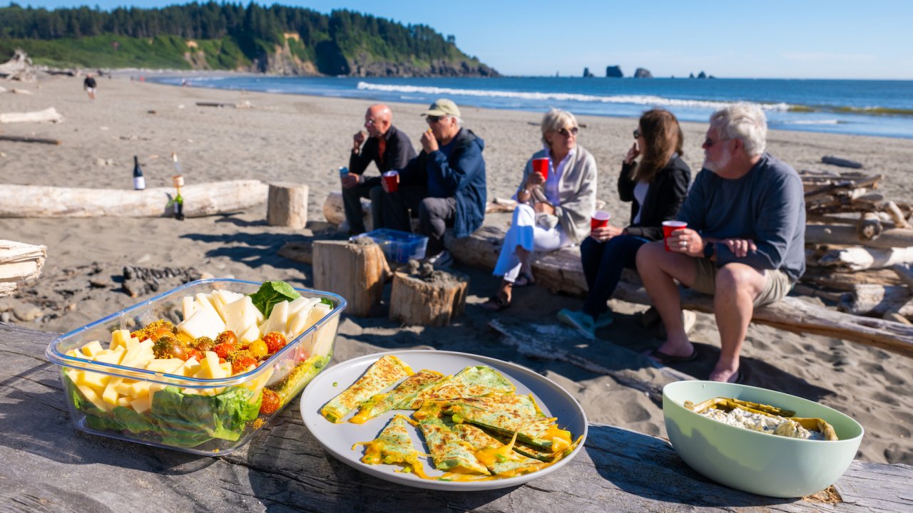 Food in Tupperware laid out on a piece of large driftwood while travelers sit and enjoy lunch on the beach in Washington state