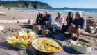 Food in Tupperware laid out on a piece of large driftwood while travelers sit and enjoy lunch on the beach in Washington state