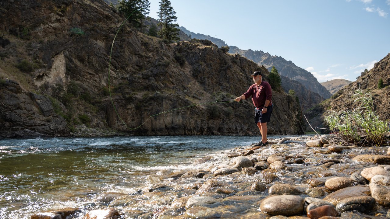A man casting a yellow fly fishing line into the Salmon River from the rocky river bank
