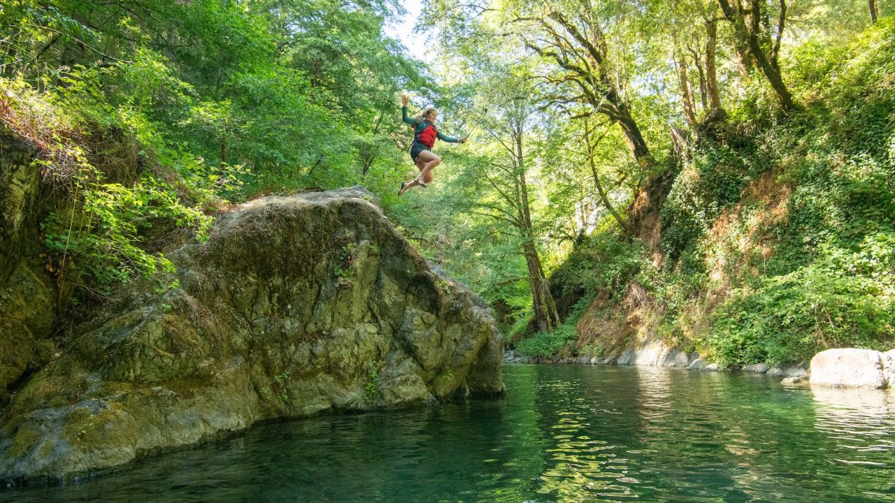 Swimming hole along the Rogue River with a person jumping off of a large rock into the pool