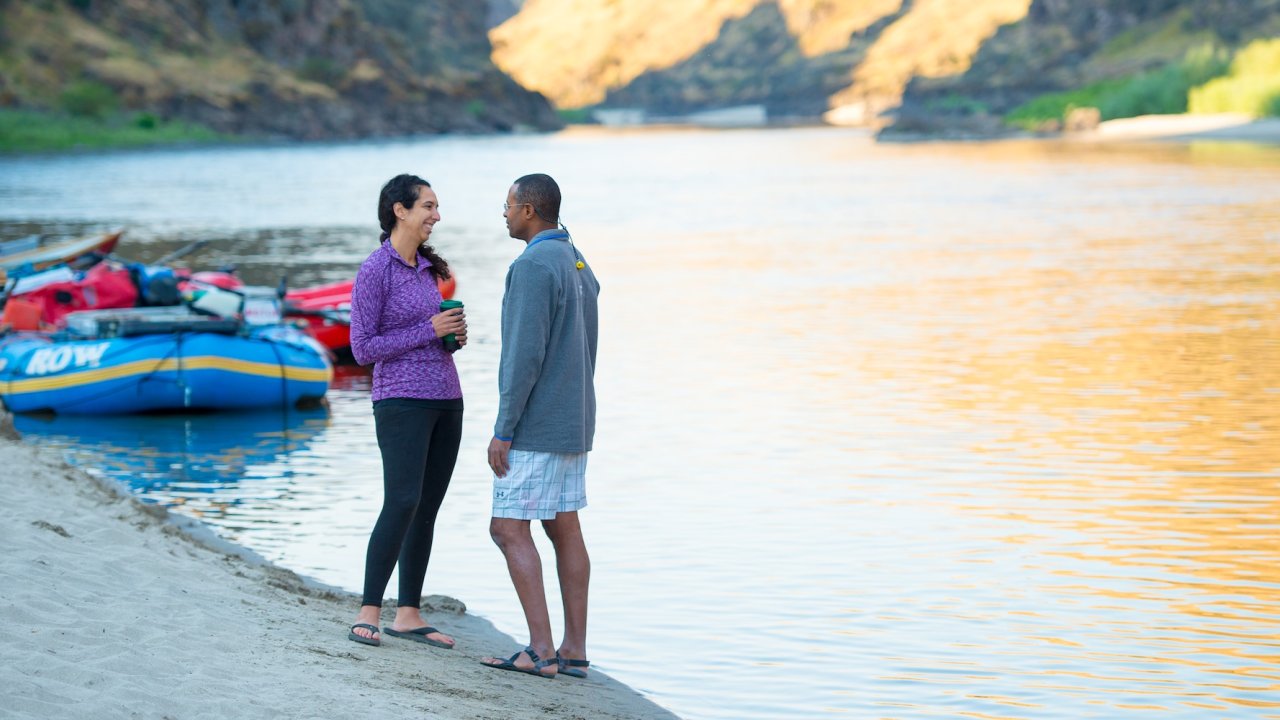 Couple standing and chatting on a sandy riverbank near colorful rafts.