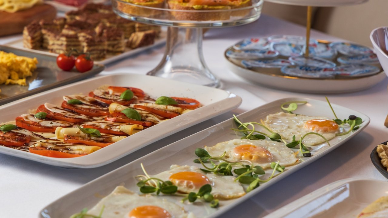 A breakfast buffet on a yacht featuring a variety of dishes on a white table cloth with a view of the sea through large windows.