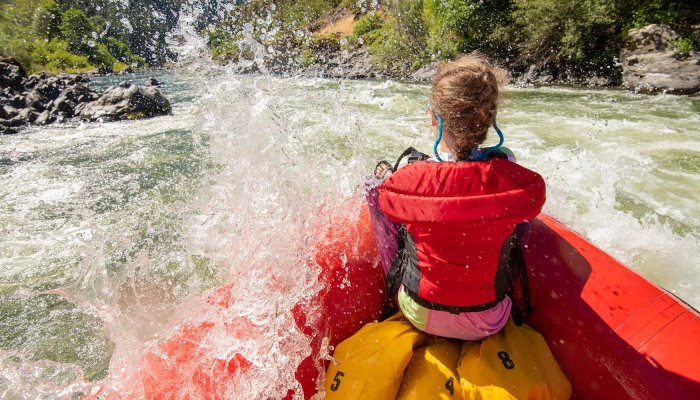 A girl in a red pft sitting at the front of a whitewater raft as it's being splashed by a wave.