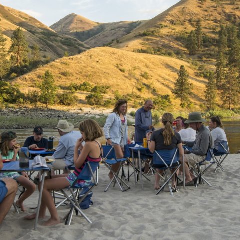 group of people sitting around tables on a sandy beach next to the river