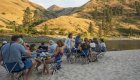 Group of people enjoying a meal on a sandy beach next to a river.
