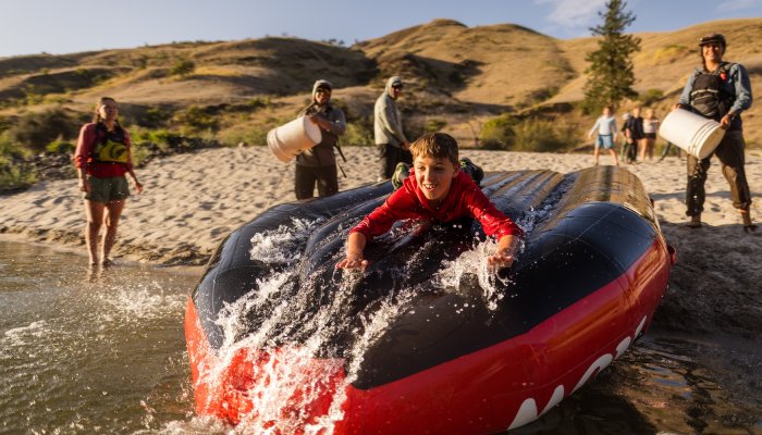 A young boy sliding down an upside down raft into the Salmon river with a sandy beach and golden mountains in the background. 