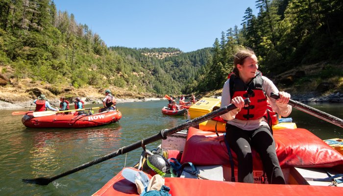 A young girl rowing a red raft down a river on a family rafting trip with multiple rafts in the background.