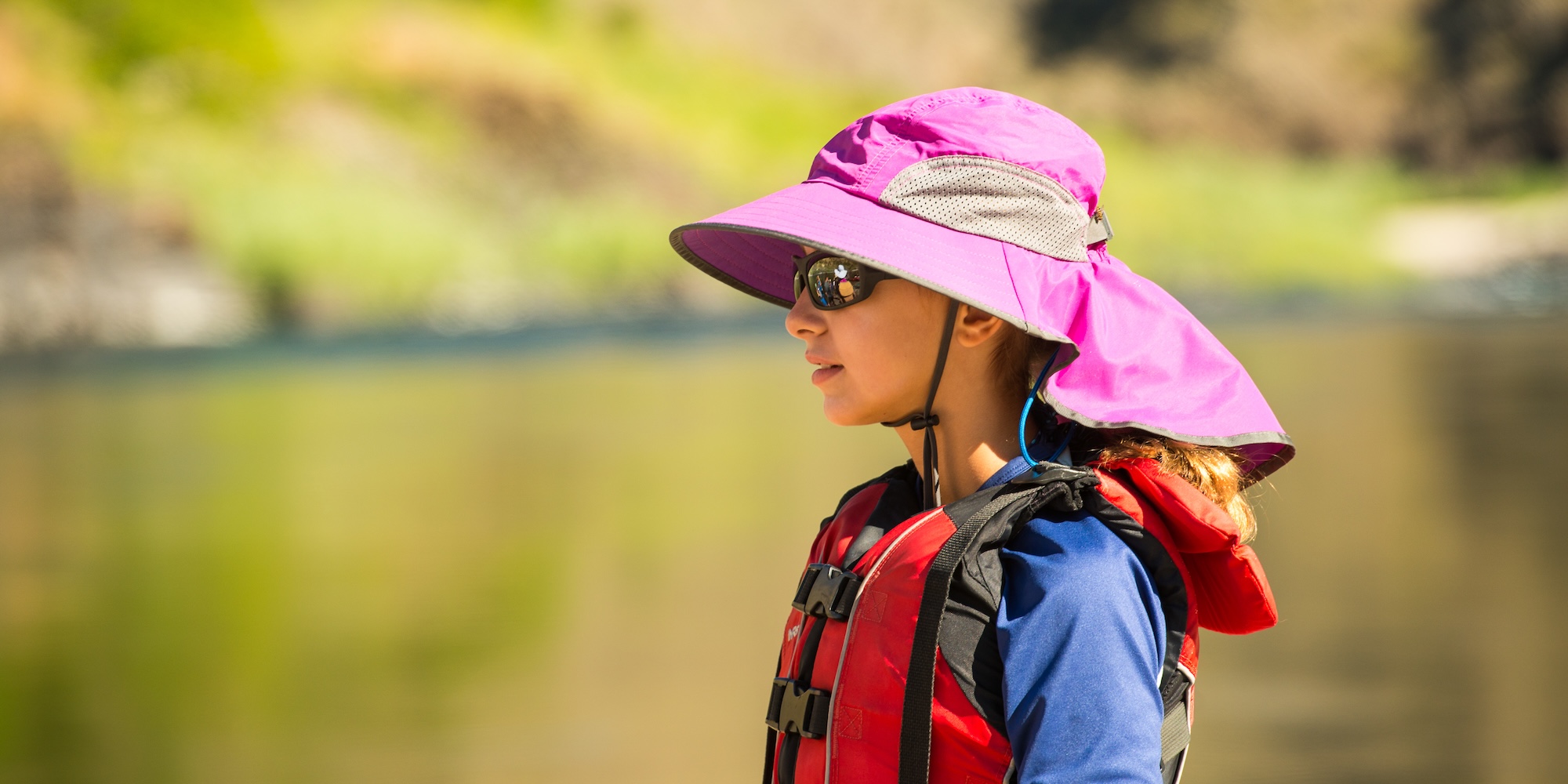 A young girl wearing a bucket hat and sunglasses next to the river. 
