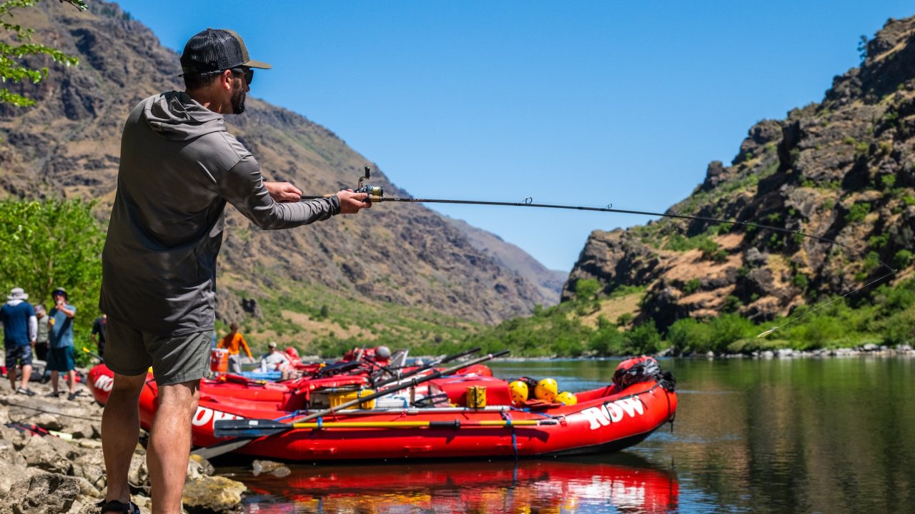 A person standing on the banks of the Snake river fishing with ROW Adventures rafts in the background.