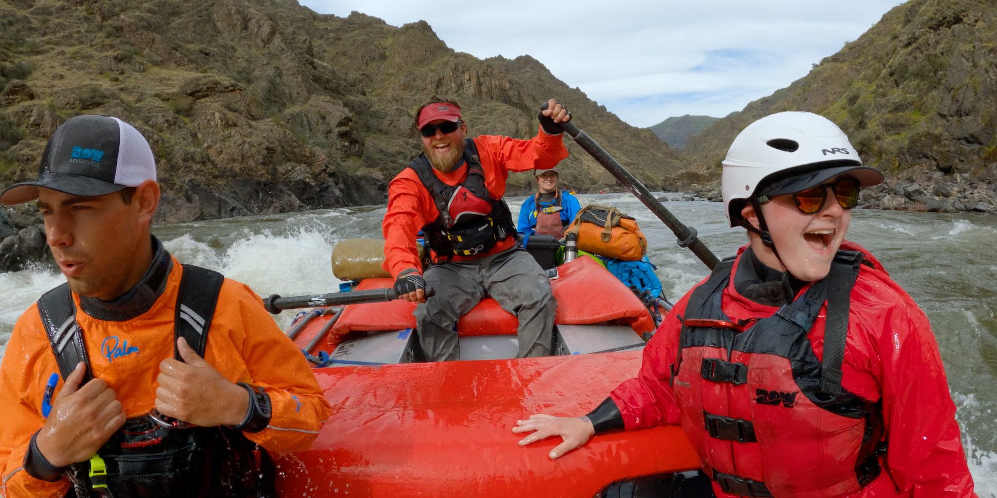 Go Pro shot from the front of a red whitewater raft looking back at the rower and passengers smiling through a rapid