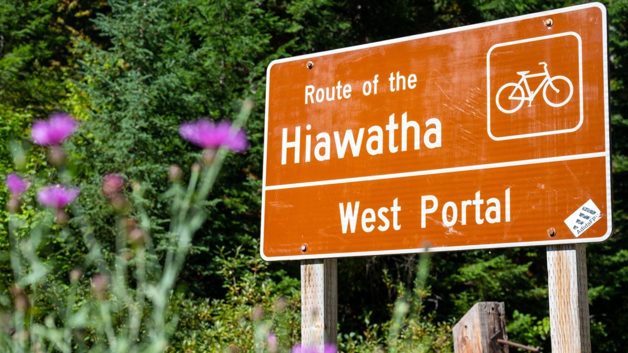 A brown trailhead sign reading 'Route of the Hiawatha – West Portal' stands beside blooming wildflowers and a lush green forest.