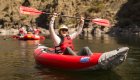 Man paddling an inflatable kayak on calm river, raising paddle in celebration.