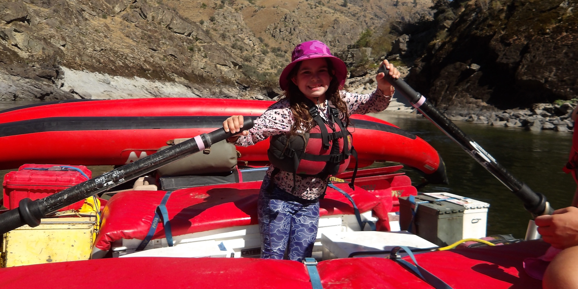 A young girl in a hat and lifejacket learning how to row a whitewater raft on a river.
