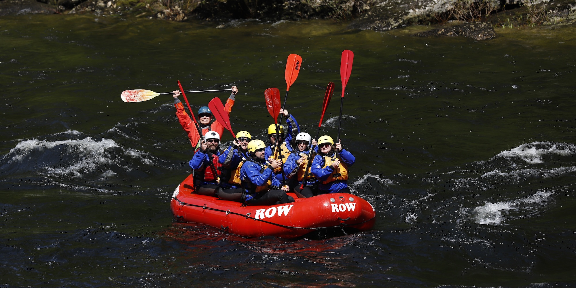 Group of people in a red ROW Adventures whitewater raft smiling and lifting their paddles up in the air. 