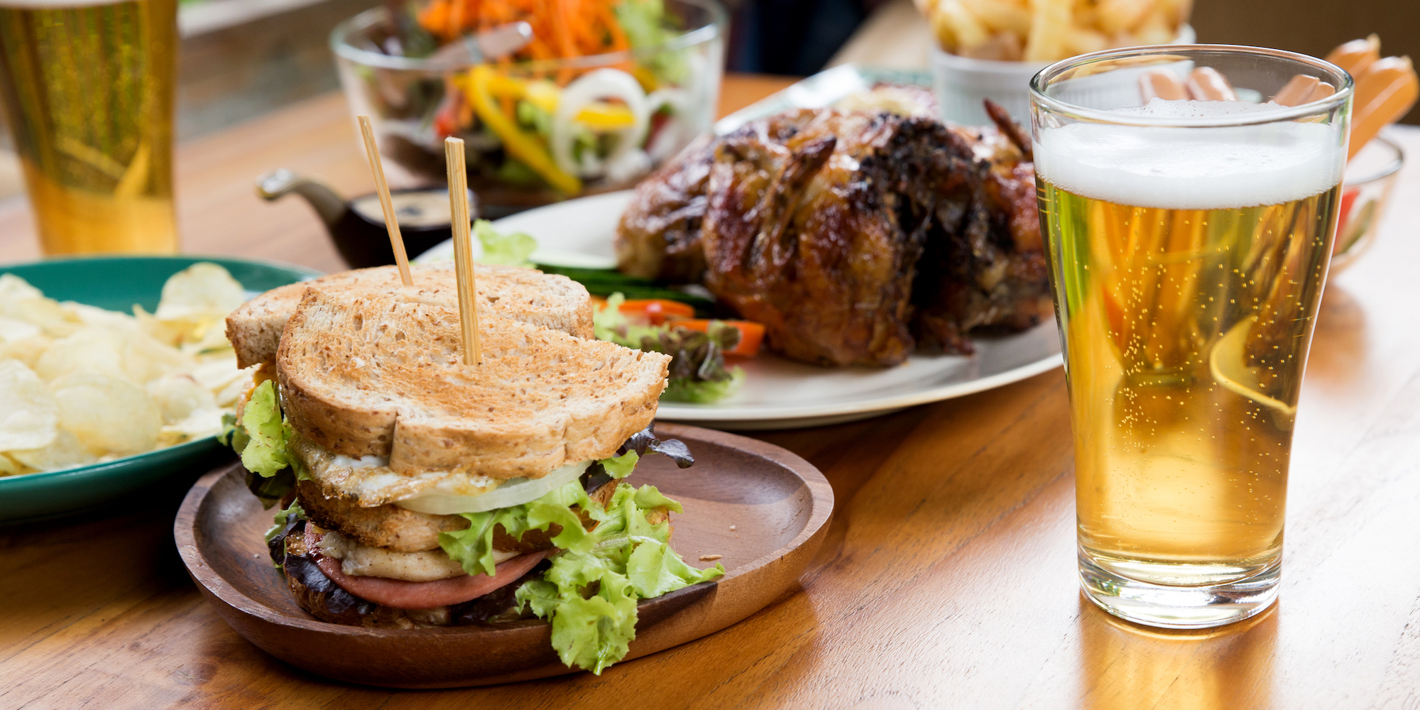 A sandwich on a brown plate next to a white plate of chicken with pints of beer spread out across the table