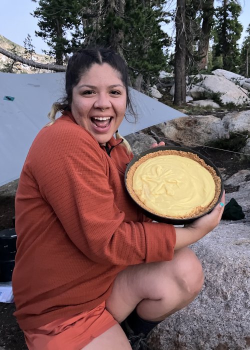 ROW Adventures employee smiling while camping and baking a cheesecake. 