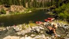 A woman with a big sun hat on sitting in a hot spring overlooking the Salmon River with red rafts tied up to shore on a sunny day