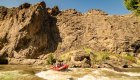 Side view of a red raft moving through a rapid with a big canyon wall behind it on a sunny day