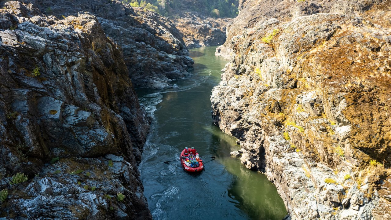 Aerial shot of a red raft floating down the Rogue River through a tight gorge with the right side in the sun and the left side of the gorge in the shade.