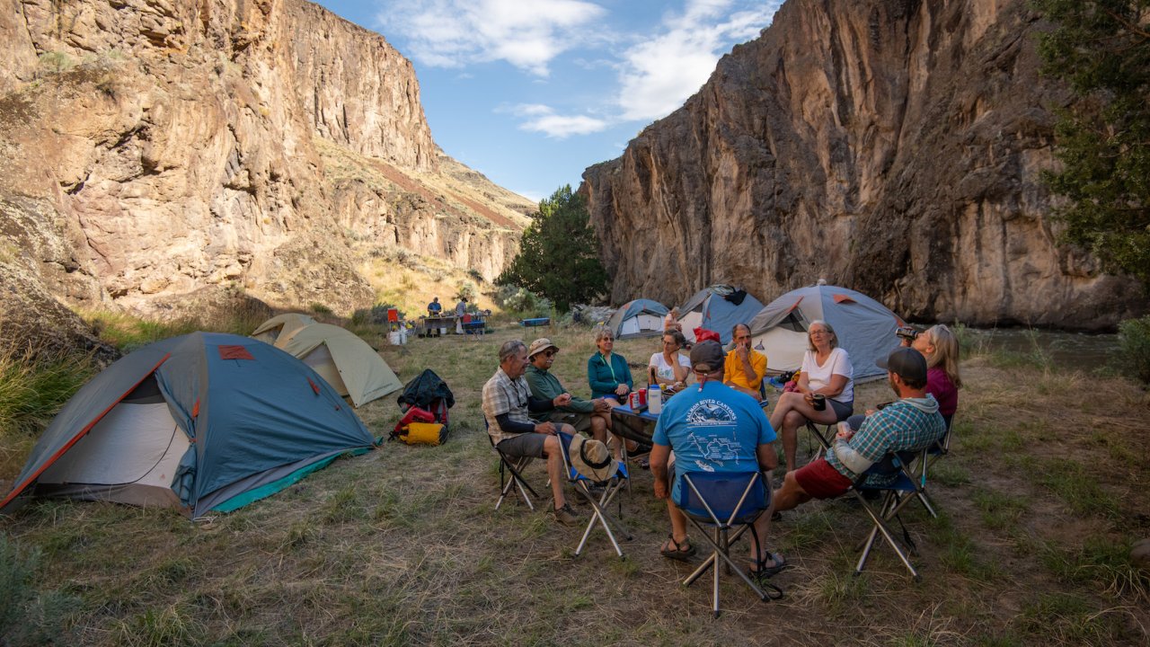 A group of people sitting around a camp table next to a group of tents at sunset