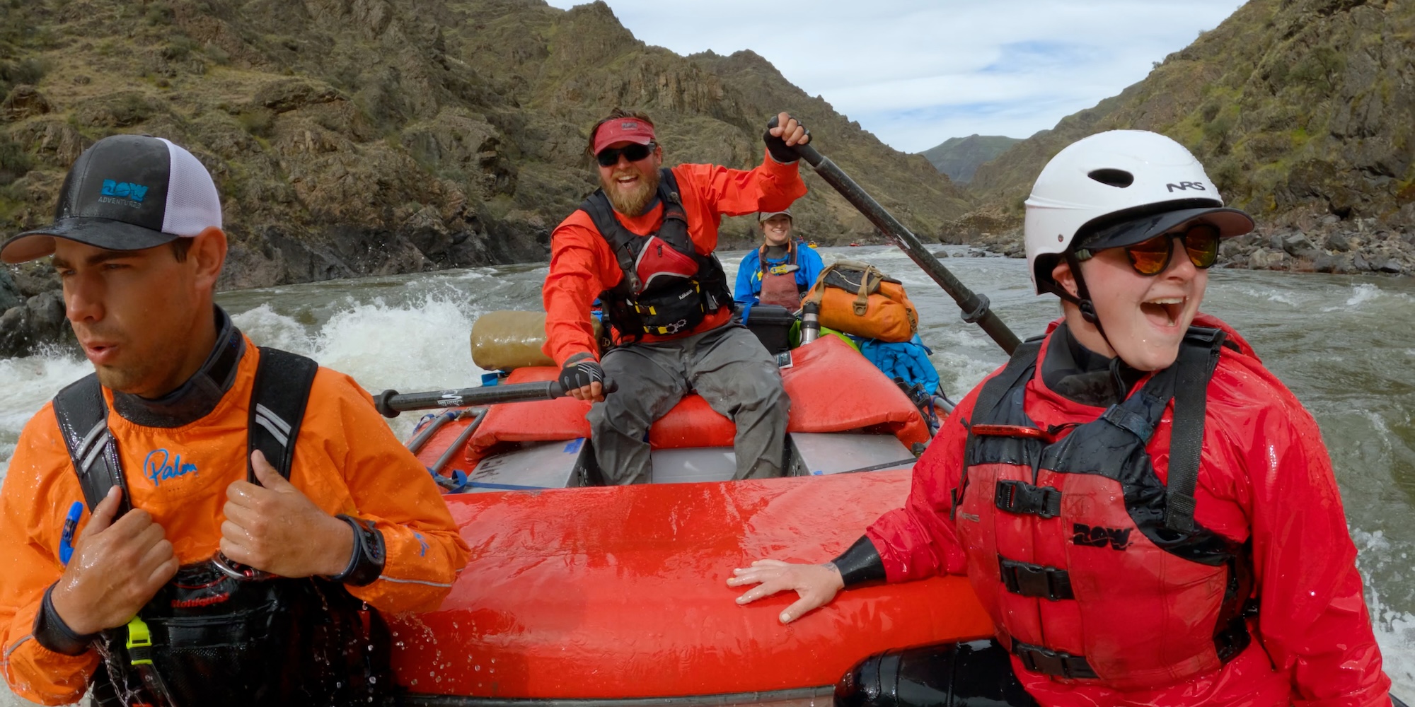 A guide grins at the oars while navigating whitewater with multiple people in the raft. 