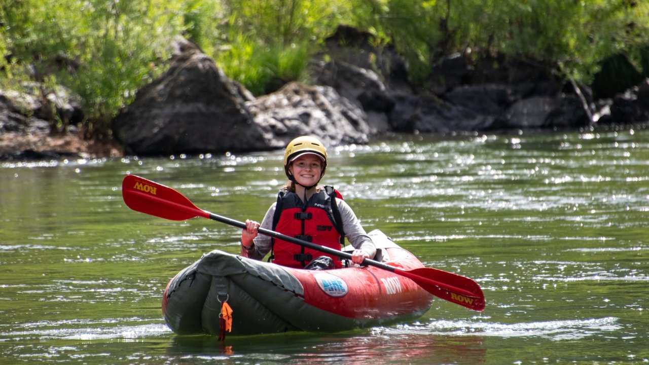 Smiling paddler in an inflatable kayak enjoying a calm stretch of the Rogue River in Oregon, a scenic spot for family-friendly Oregon rafting trips.