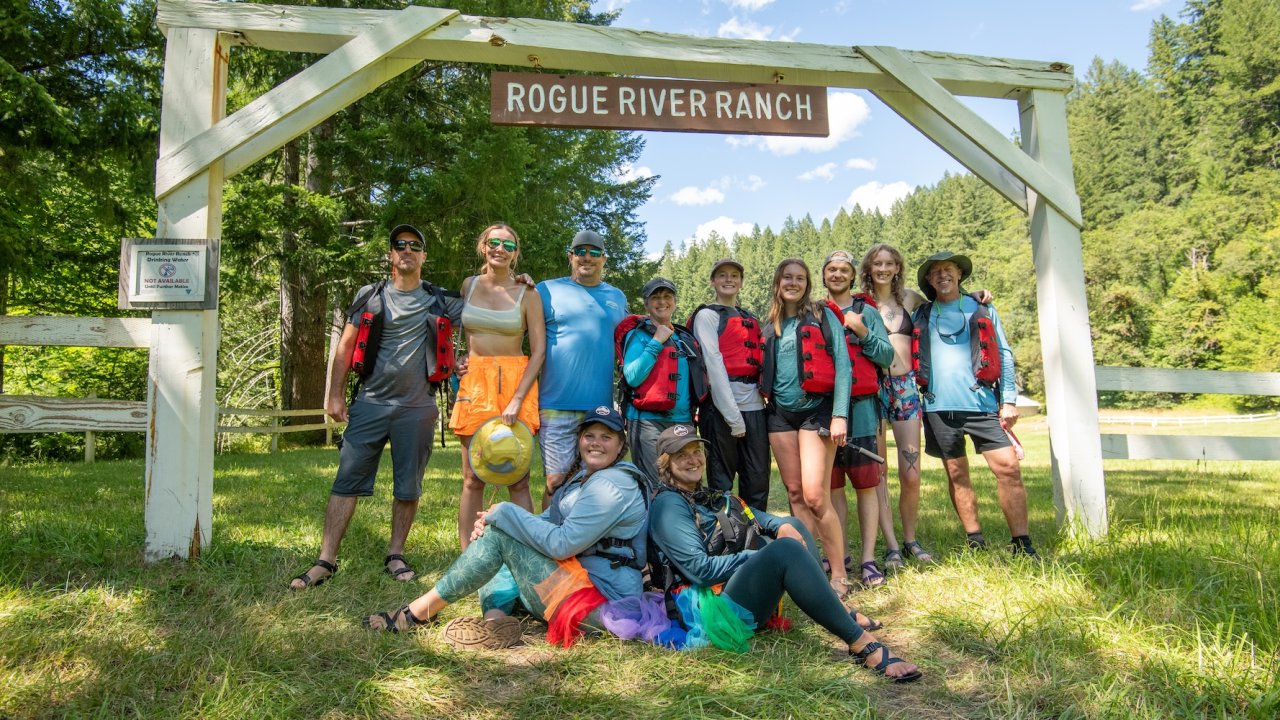 Group of whitewater rafters sitting under a sign at a lodge next to the river