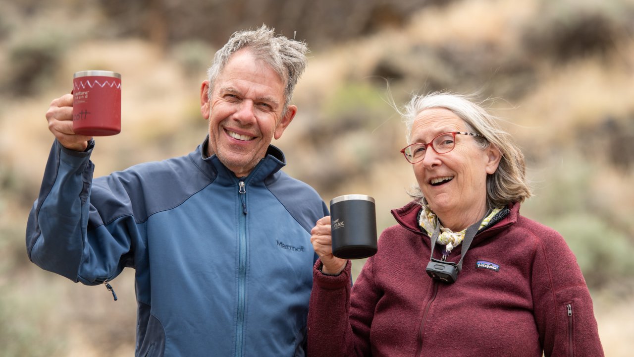 Close up on a man on the left and woman on the right holding up aluminum camping mugs and smiling