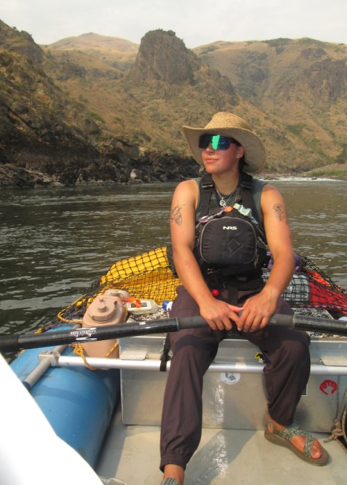ROW Adventures river guide sitting in a raft rowing down a river with mountains and rapids behind them. 