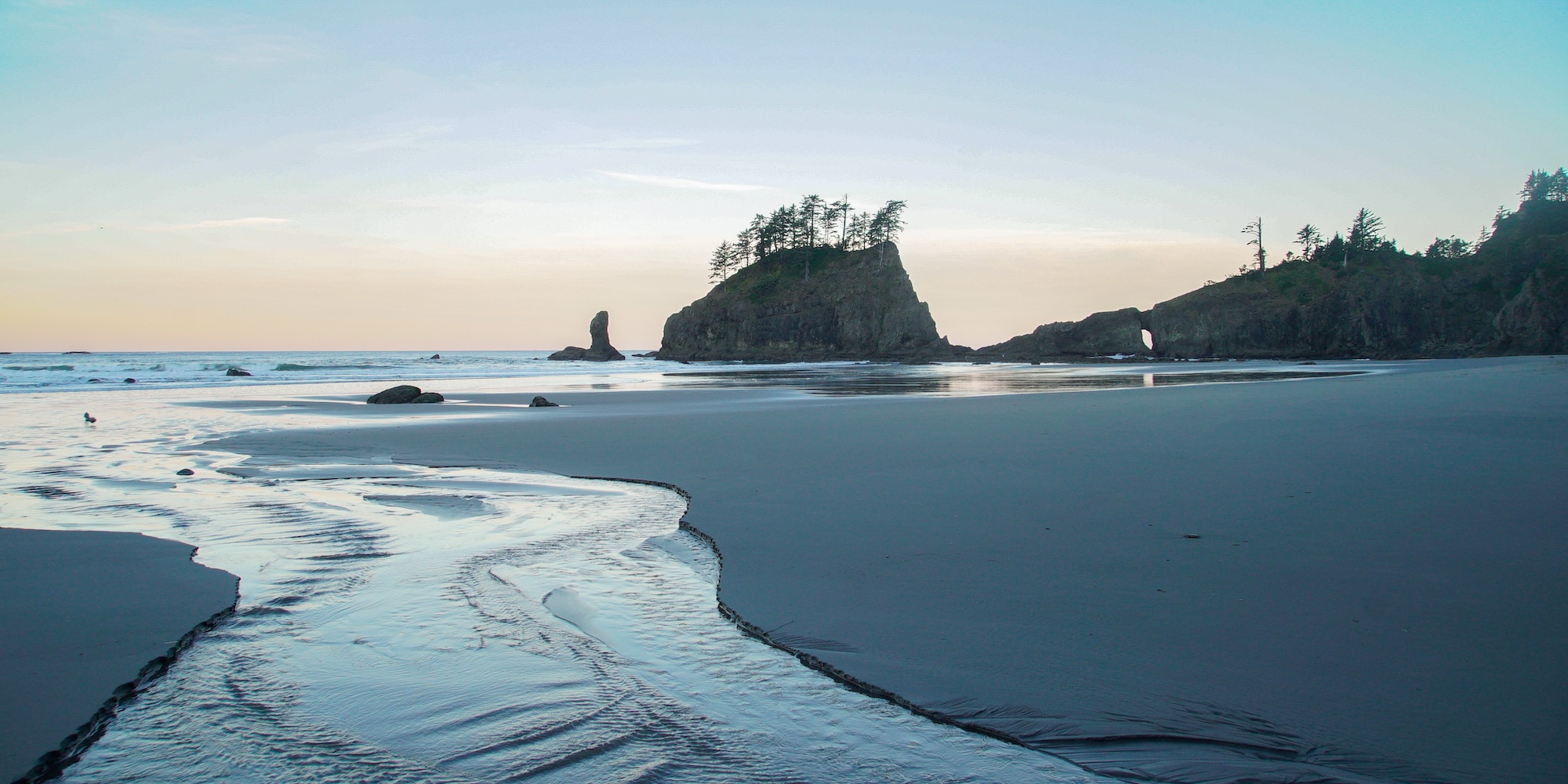 A peaceful sunrise over sea stacks at Second Beach on the Olympic Peninsula, where ocean waves meet smooth sand and rugged rock formations.