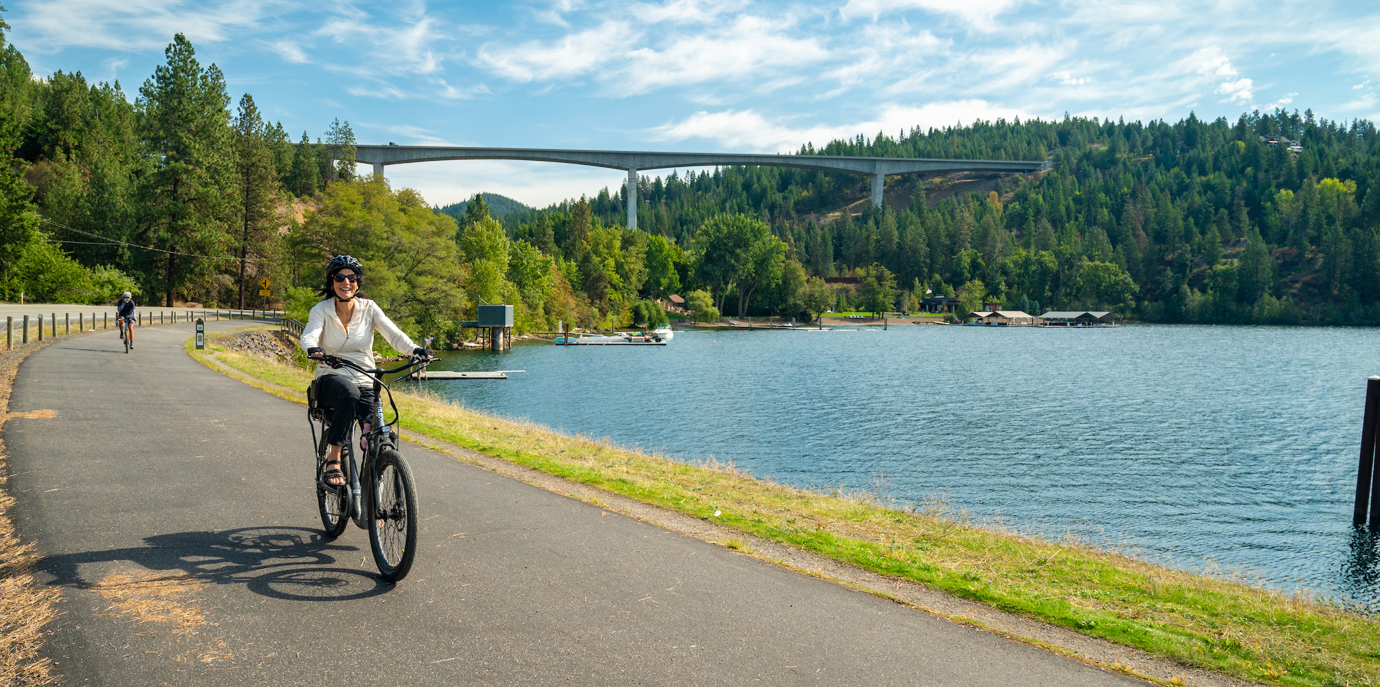 Two people biking and smiling while biking towards the camera along the Trail of the Coeur d'Alenes in North Idaho