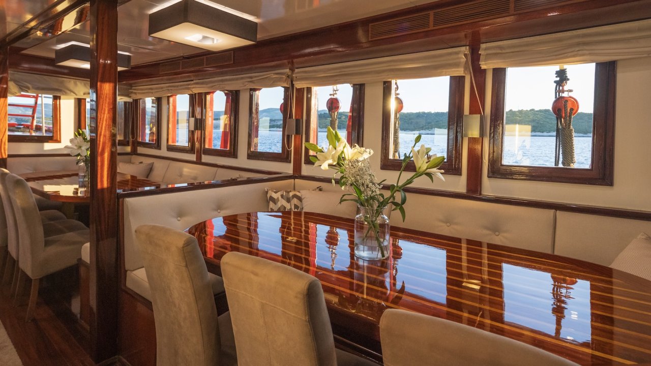 A yacht's indoor dining area with a polished wooden table, cushioned seating, and windows offering a view of the Adriatic Sea.