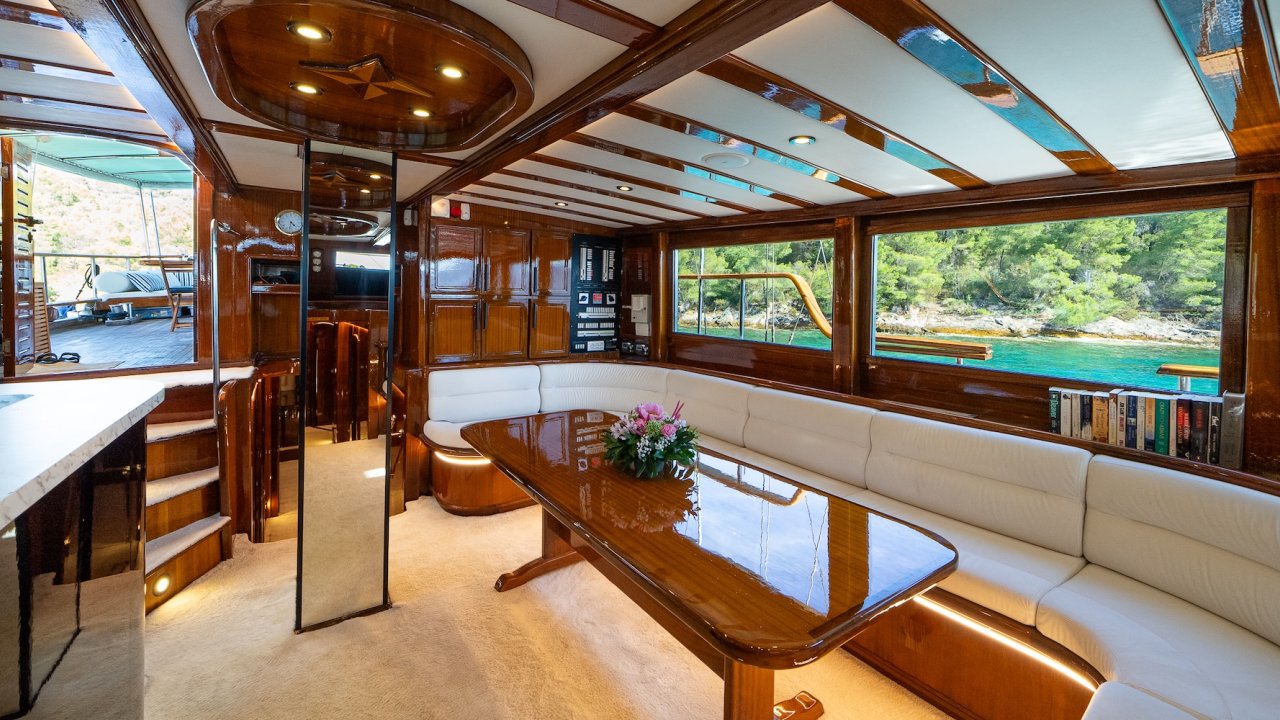 A yacht salon with polished wood interiors, long seating area, and large windows.